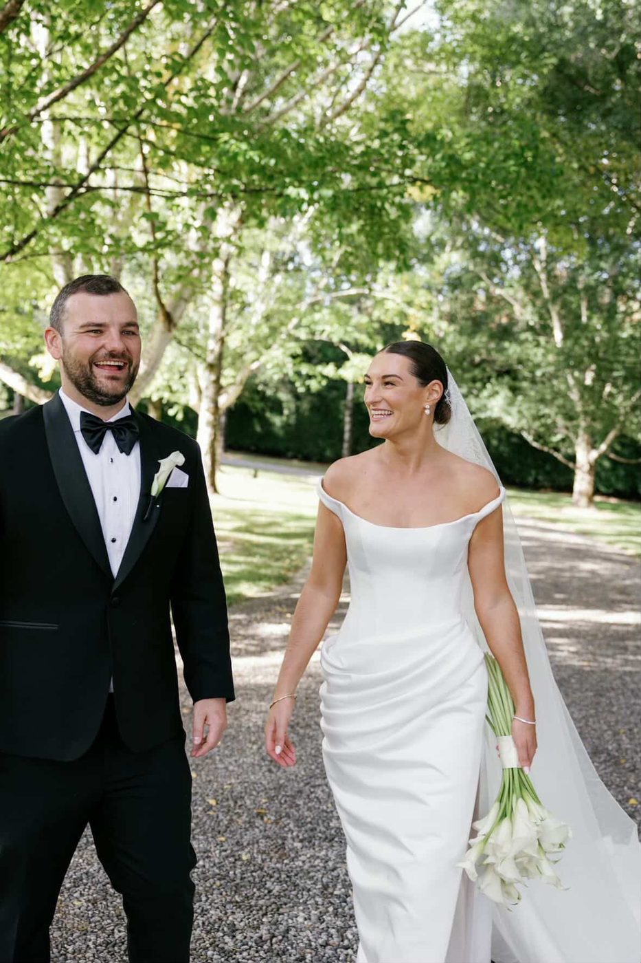 Bride and groom walking down a tree-lined path, her calla lilies in hand