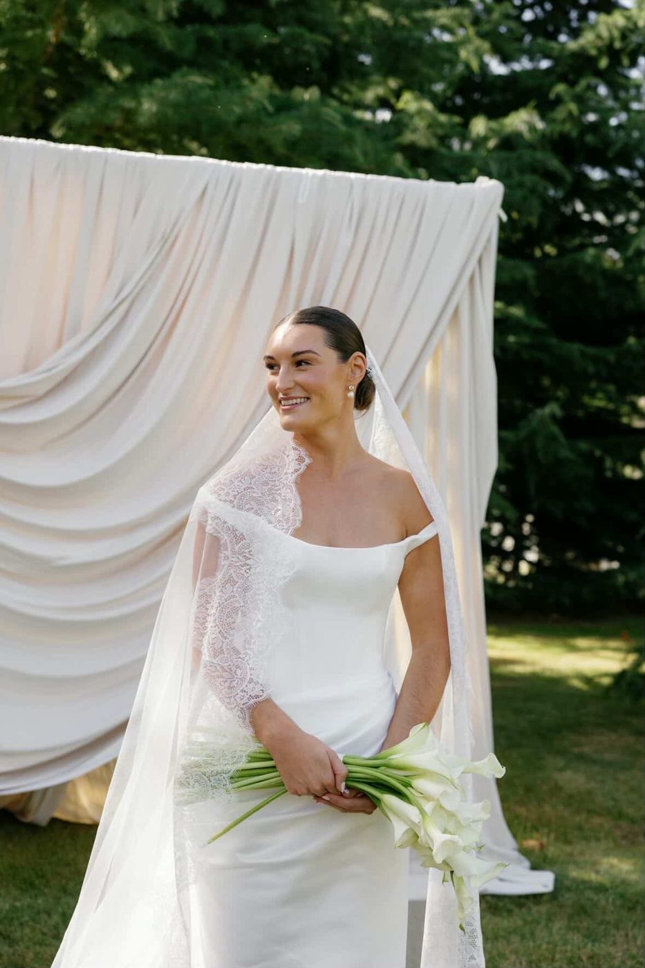 Bride in an off-the-shoulder gown and lace-trimmed veil holding white calla lilies